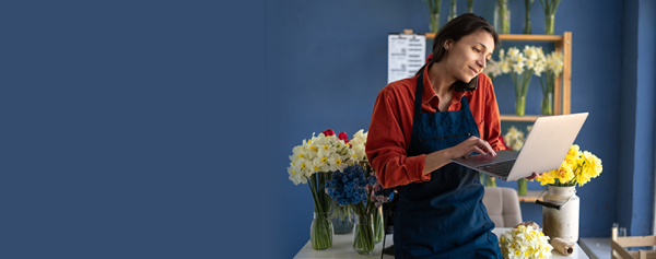 Florist wearing a blue apron uses a laptop in her flower shop, surrounded by colorful bouquets and floral arrangements.