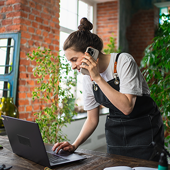 A person surrounded by plants talks on their smartphone while looking down into a laptop screen.