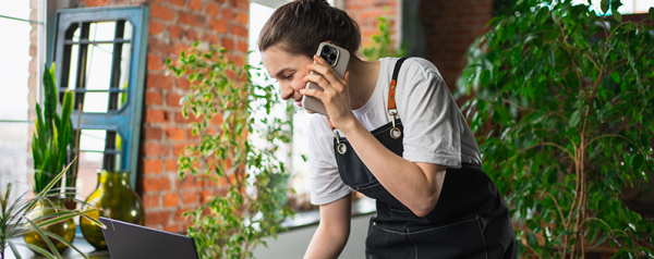 A person surrounded by plants talks on their smartphone while looking down into a laptop screen.