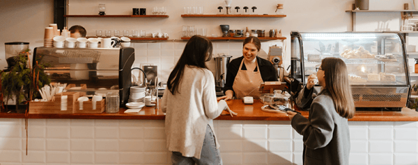 An employee behind the counter at a modern coffee shop helps two customers.