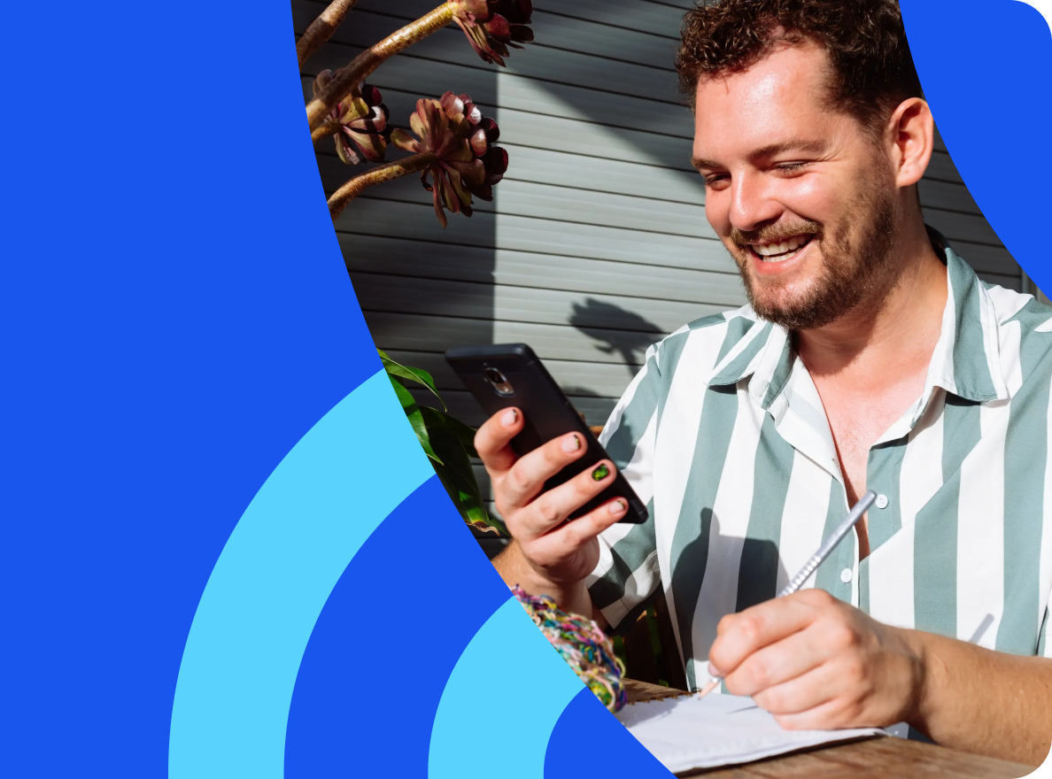 Bearded man writing notes in a notebook while holding a smartphone outdoors